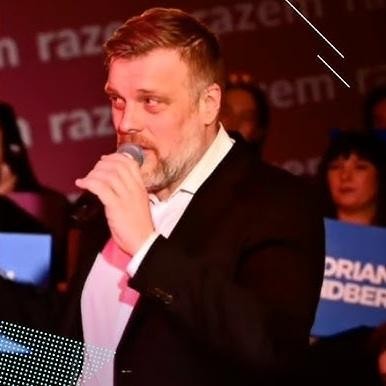 <alt>Adrian Zandberg przemawiający z energią na wiecu politycznym, gestykulujący, z rozmytym tłem sugerującym tłum i wydarzenie publiczne.</alt>
