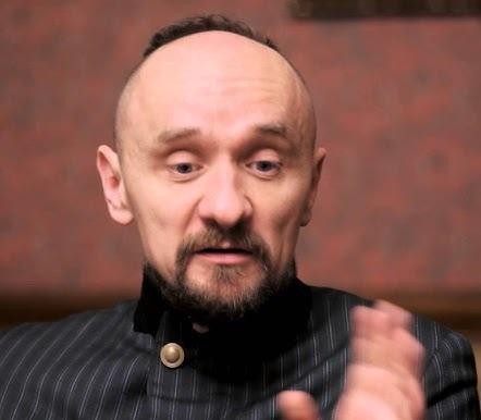 Jan Klata, reżyser, dynamicznie kierujący próbą teatralną, z aktorami i elementami scenografii w tle.