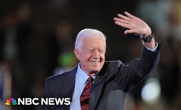 Jimmy Carter w późnym wieku, aktywnie pracuje przy budowie domu dla Habitat for Humanity, ubrany w koszulę roboczą i okulary ochronne, wbijający gwoździe w drewnianą ramę.