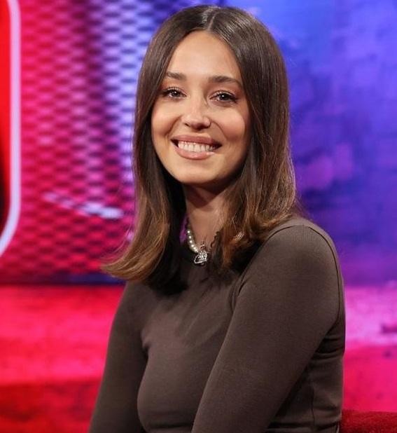 Izabella Krzan jako prezenterka telewizyjna w studio, uśmiechnięta i dynamiczna, z mikrofonem w dłoni, oddająca energię i charyzmę swojej pracy.