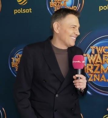 Krzysztof Ibisz w studiu telewizyjnym, dynamicznie prowadzący program, otoczony światłami studyjnymi, w eleganckim garniturze.